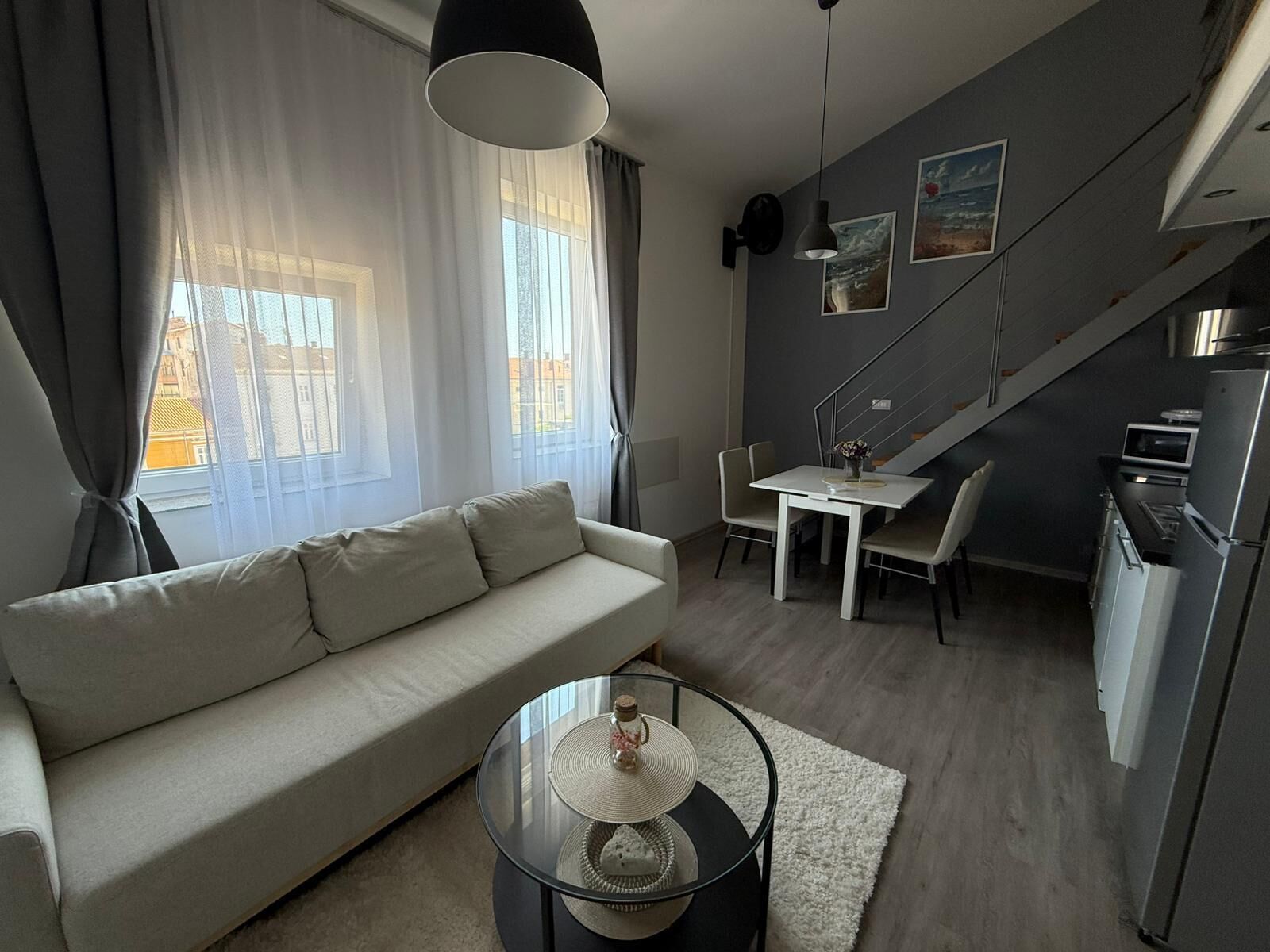 A modern living space with a sofa, dining table, chairs, a glass coffee table, and a kitchen area with a refrigerator and microwave.