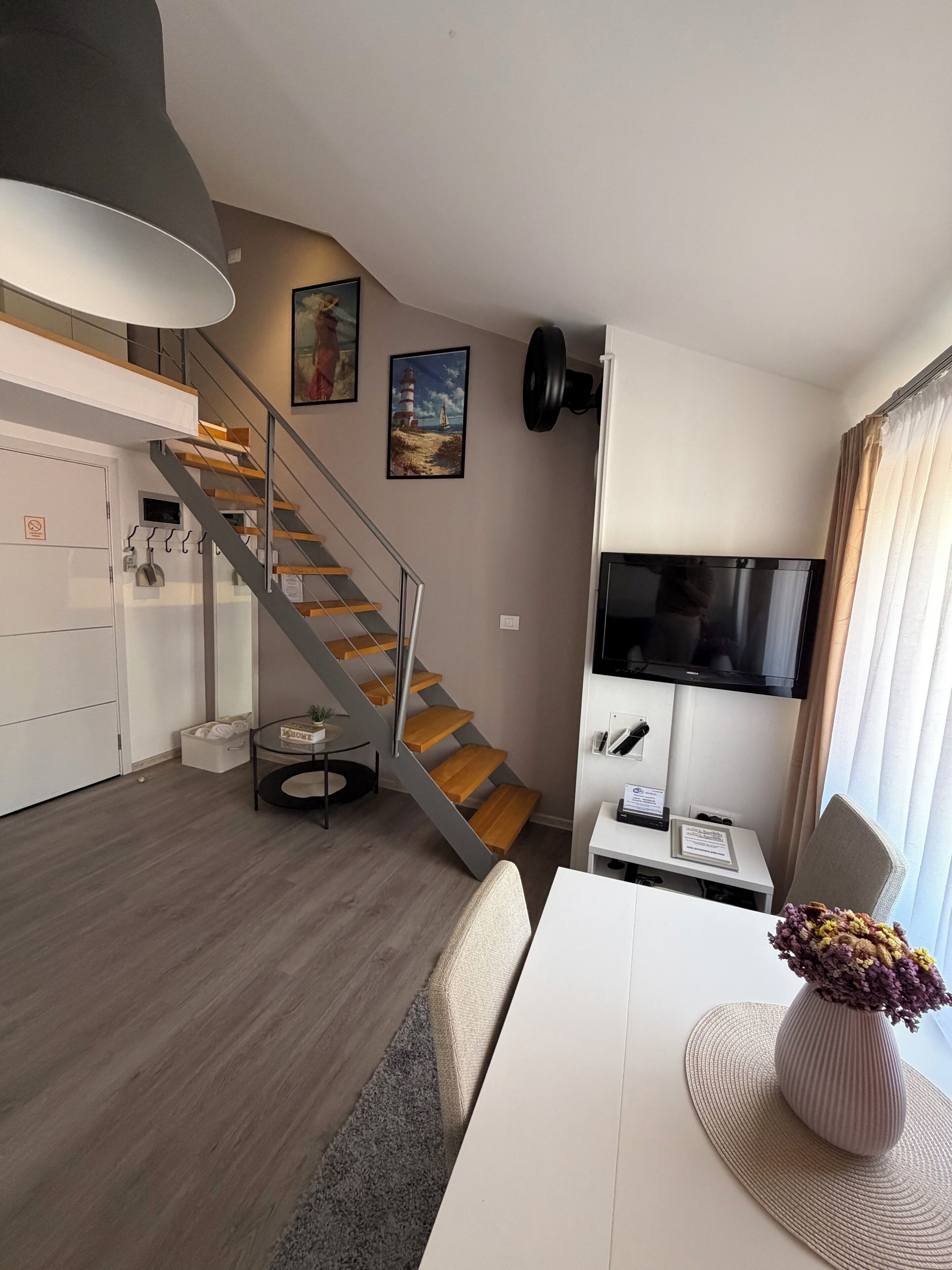 Een moderne woonruimte met een trap, een flatscreen televisie, een eethoek en een kleine tafel met een vaas.