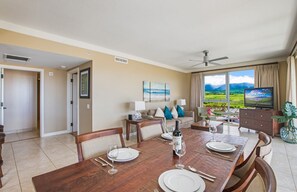 Un coin repas avec une table en bois dressée pour quatre personnes, un téléviseur et une vue panoramique sur les montagnes, visible à travers de grandes fenêtres.