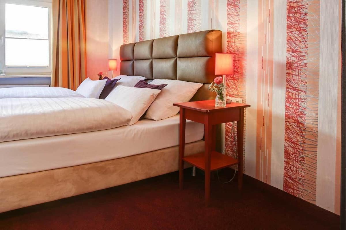 A double bed with a wooden frame and a headboard, a bedside table with a lamp, and a window with curtains.
