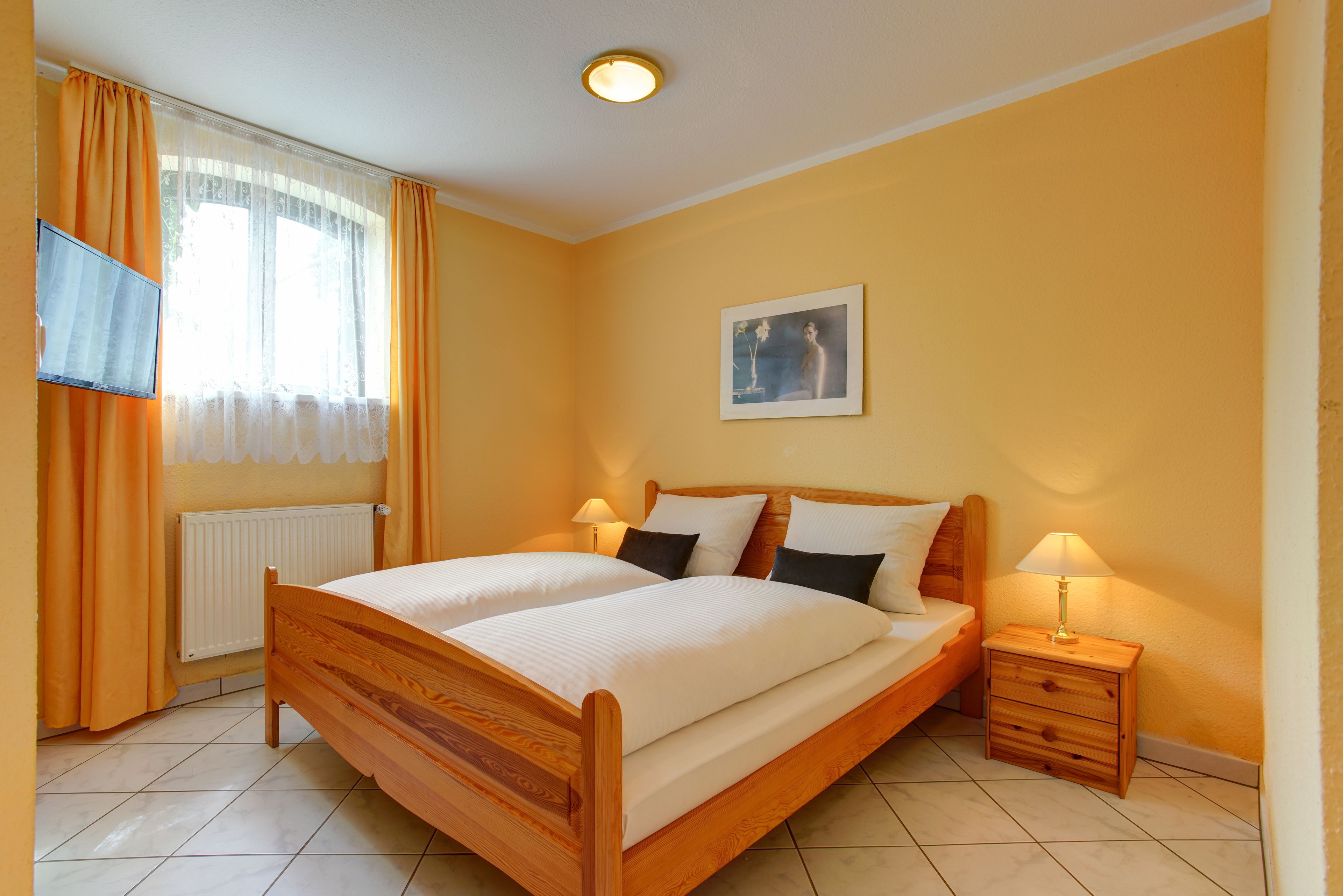Un dormitorio con cama de madera, dos mesitas de noche, televisor en la pared y ventana con cortinas.