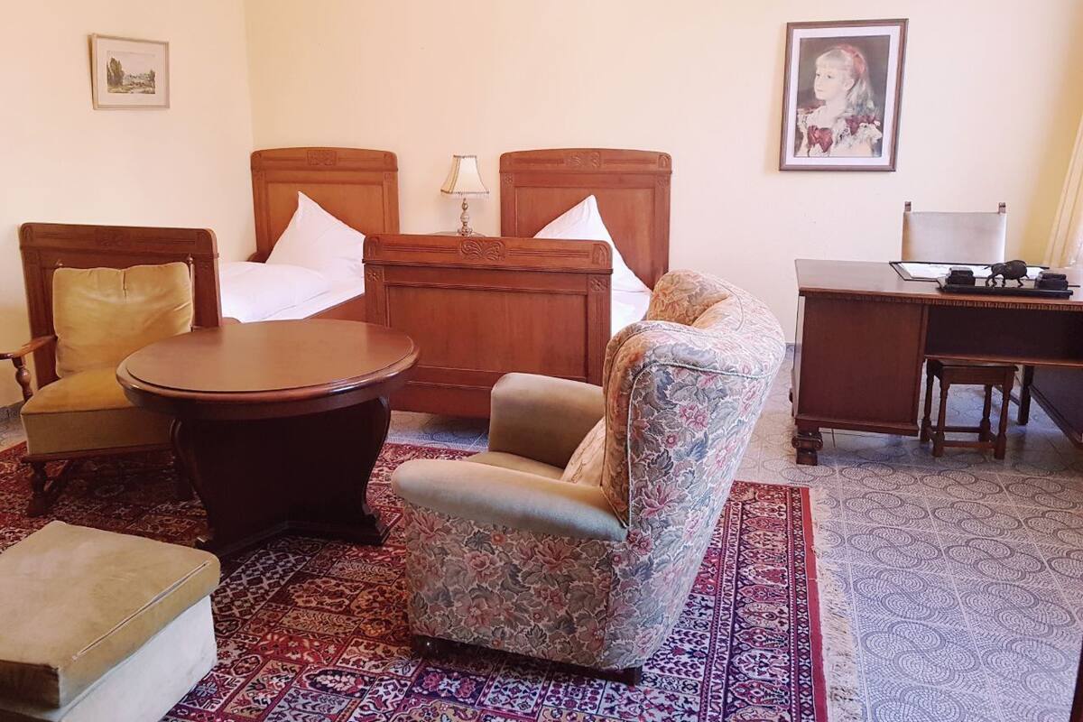 A room with a wooden bed, a round wooden table, a floral armchair, a desk, and a chandelier.
