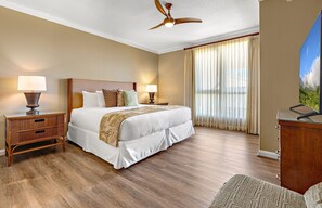 A hotel room with a bed, two bedside tables with lamps, a ceiling fan, a large window with curtains, and a television on a stand.