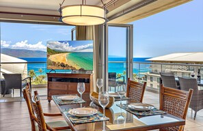 A dining area with a table set for two, a television displaying a beach scene, and a view of the ocean.