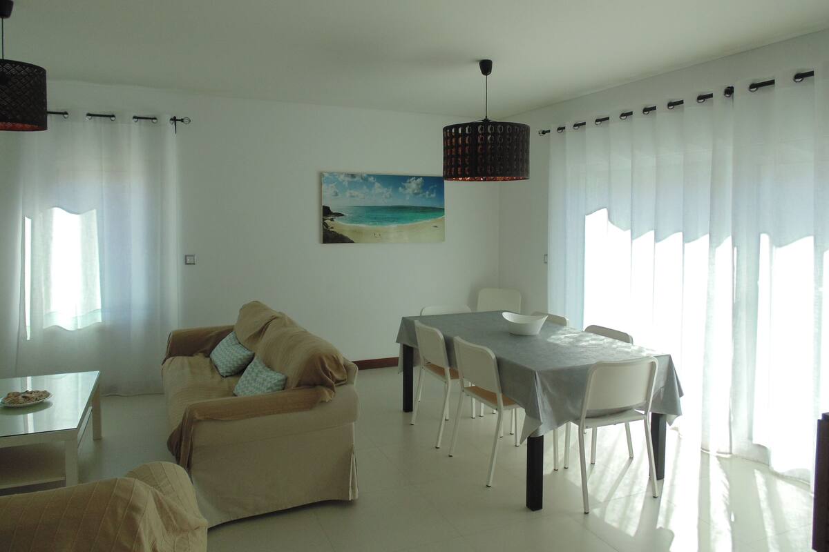 A living room with a sofa, dining table, chairs, and a painting of a beach.