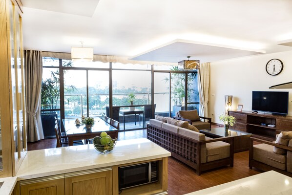 A modern living room with a sofa set, a dining table with a bowl of fruit, a TV, and a balcony with a view of the city.