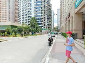 A street view with a row of high-rise residential buildings, a sidewalk, and a few potted plants along the curb.