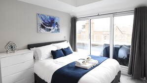 A modern hotel room with a large bed, a bedside table with a clock, a painting on the wall, and a view of the city through the window. - Northside Apartments Ealing (London)