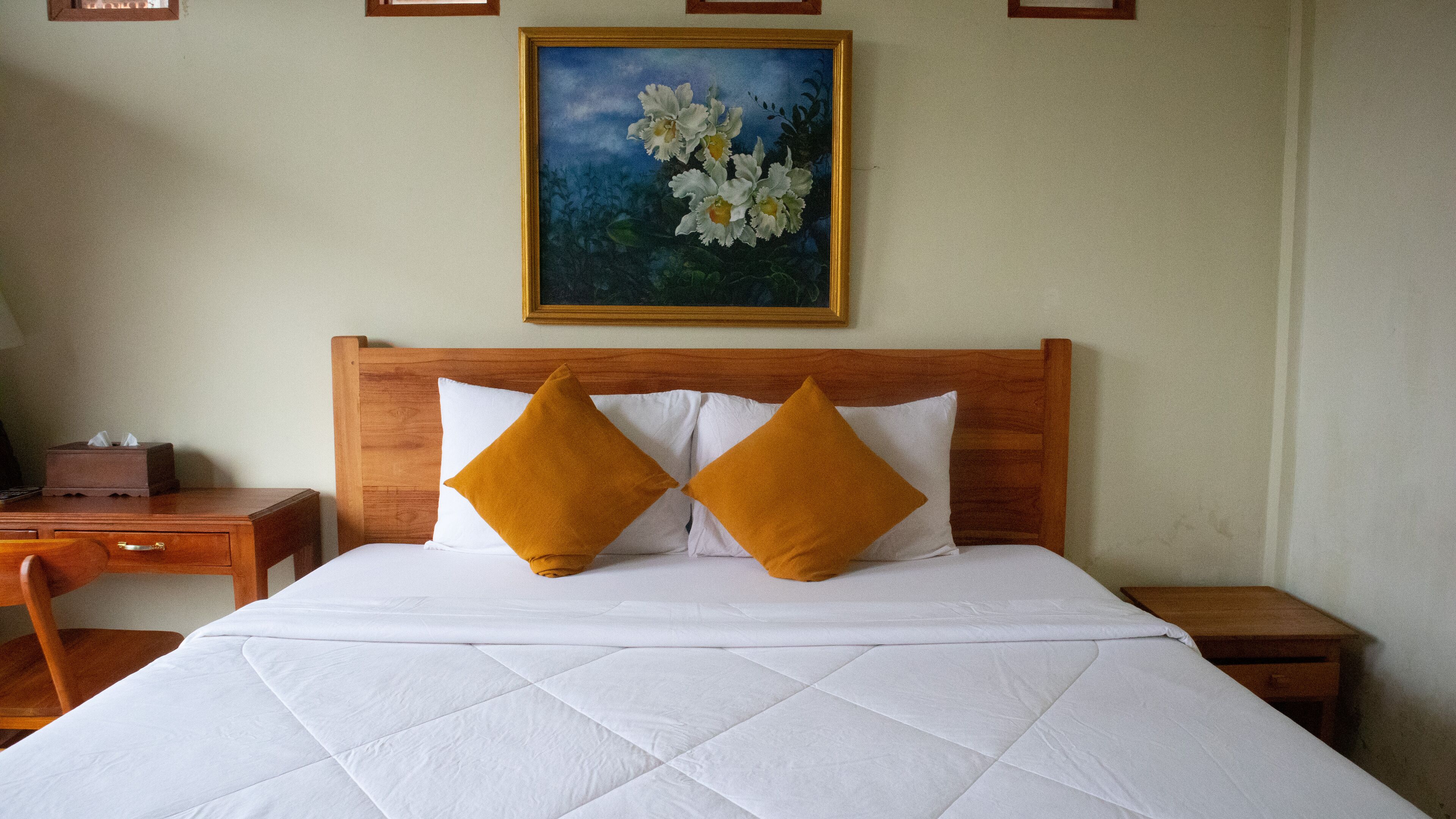 Una cama bien hecha con un cabecero de madera, dos almohadas blancas y dos almohadas color mostaza. Un cuadro de flores cuelga sobre la cama.