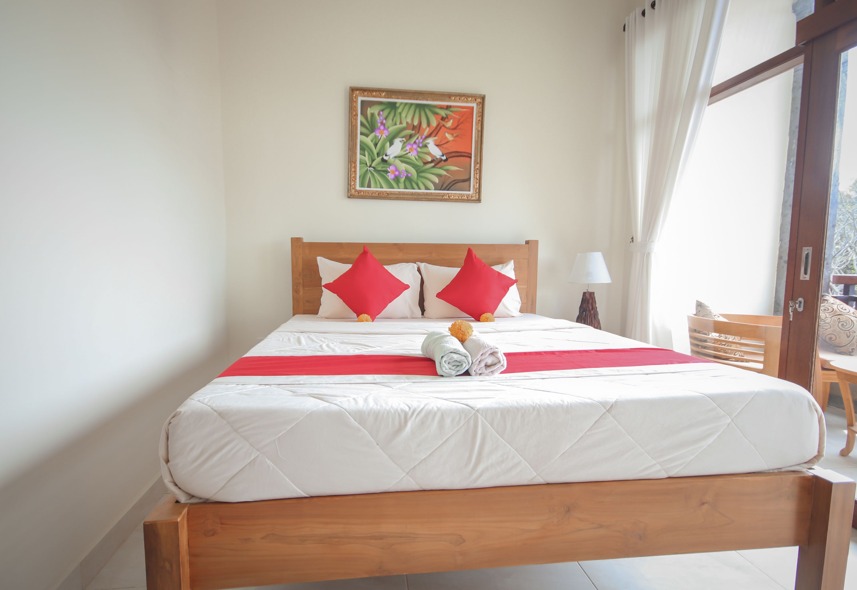A neatly made bed with a wooden headboard, red pillows, and a rolled towel. A painting hangs on the wall above the bed.