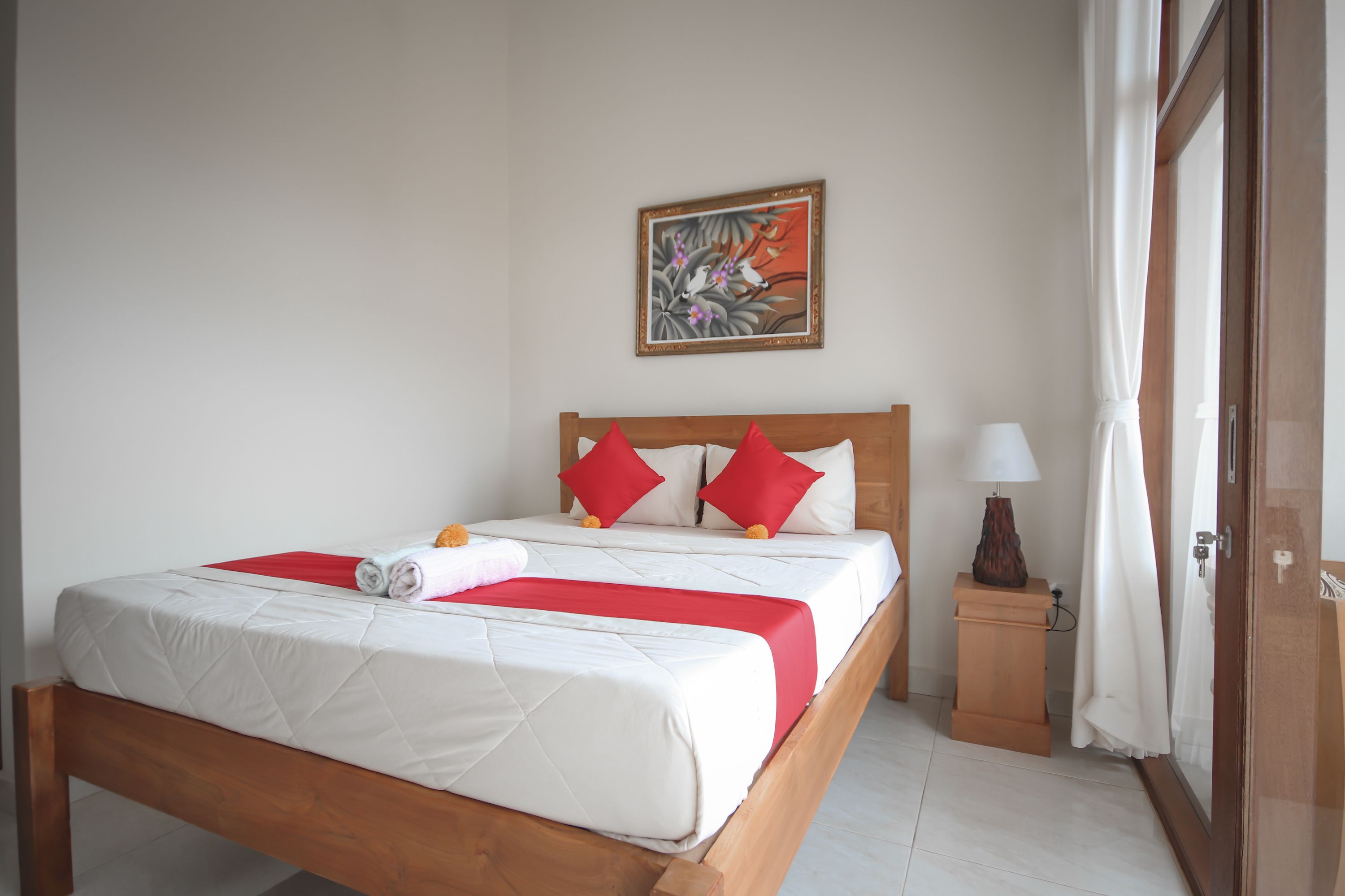 A neatly made bed with red and white bedding, a wooden headboard, a small bedside table with a lamp, and a painting on the wall.