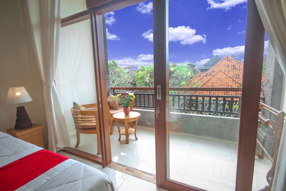 Habitación de hotel con cama, mesita de noche con lámpara, zona de comedor con mesa y sillas, y puerta corrediza de vidrio que da a un balcón con vistas a edificios y árboles.
