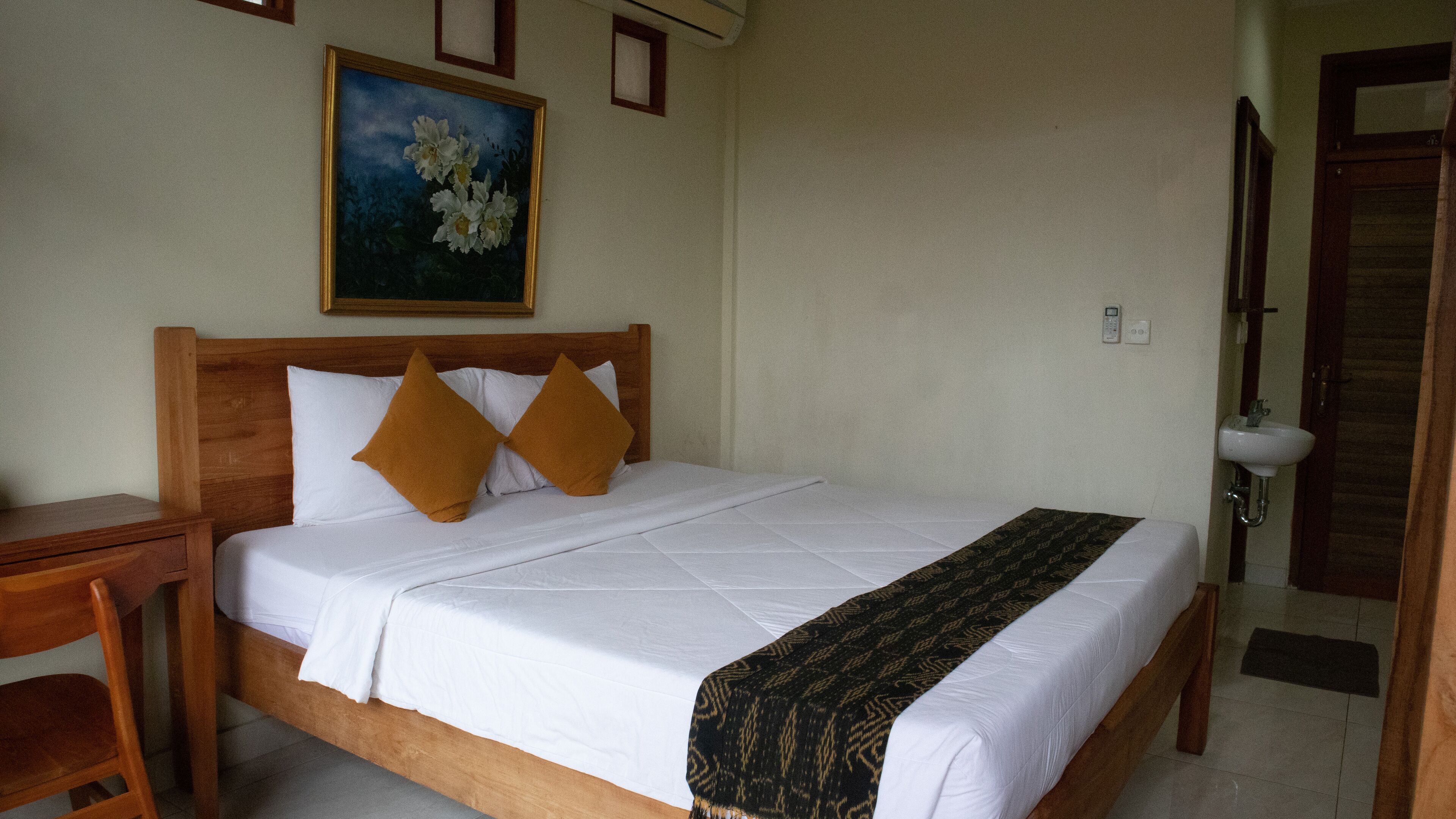 Una cama bien hecha con sábanas blancas y dos almohadas marrones, una mesita de noche de madera con una lámpara, un cuadro de flores en la pared, y un baño con un lavabo y un inodoro a la vista.