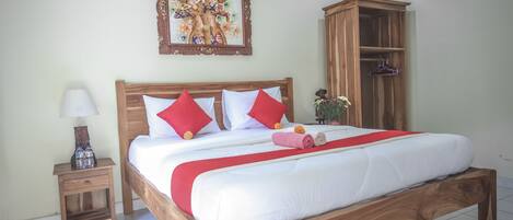 A neatly made bed with a wooden frame, white bedding, and red accents. There is a small nightstand with a lamp and a decorative item. A wooden wardrobe is against the wall, and a framed picture hangs above the bed.