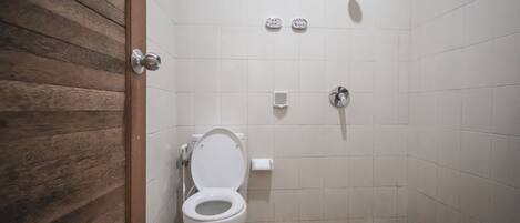 Une salle de bain avec une porte en bois, des toilettes blanches, une douche et un sol carrelé.