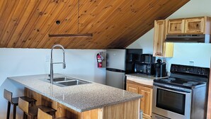 A modern kitchen with a wooden ceiling, granite countertop, stainless steel appliances, and wooden bar stools. - Sara Placid Inn & Suites (Saranac Lake)