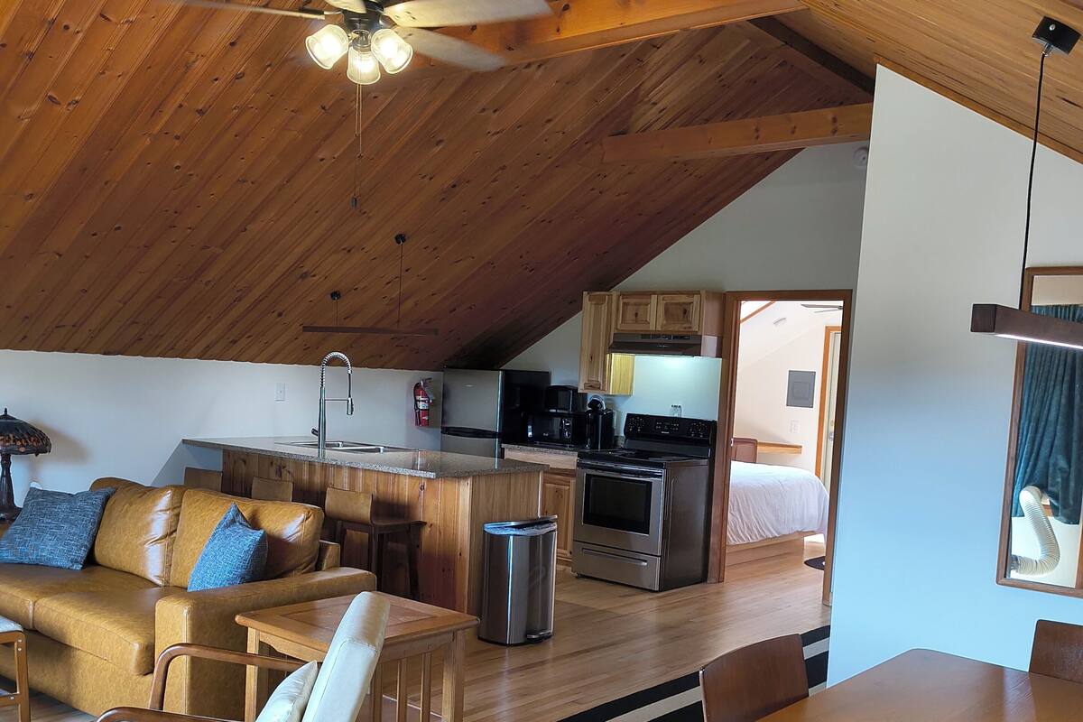 A cozy loft-style living space with a wooden ceiling, a dining area, a kitchen, and a visible bedroom.