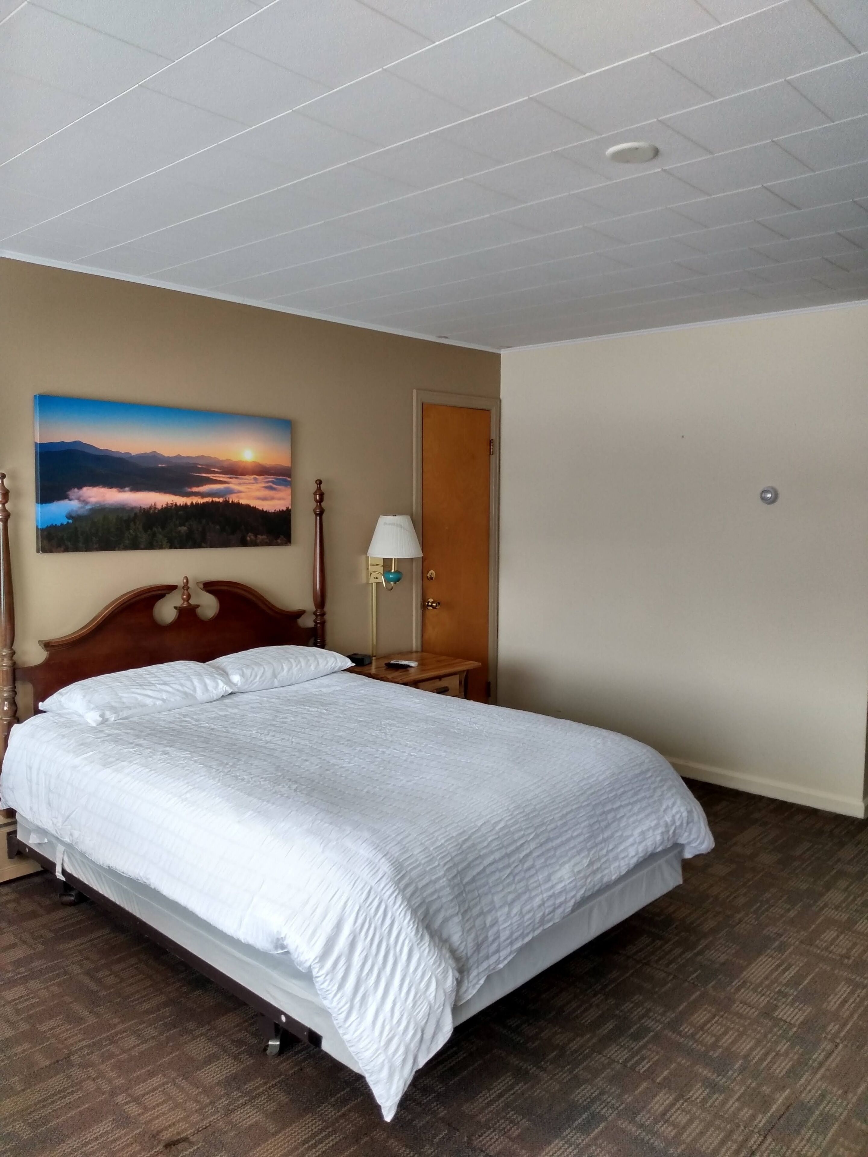 Habitación de hotel con una cama grande, cabecera de madera, mesita de noche, lámpara, puerta y un cuadro de un atardecer sobre montañas.