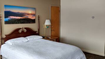 Habitación de hotel con una cama grande, cabecera de madera, mesita de noche, lámpara, puerta y un cuadro de un atardecer sobre montañas.