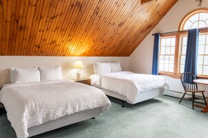 A room with two beds, a wooden ceiling, a window with curtains, and a small table with a chair.