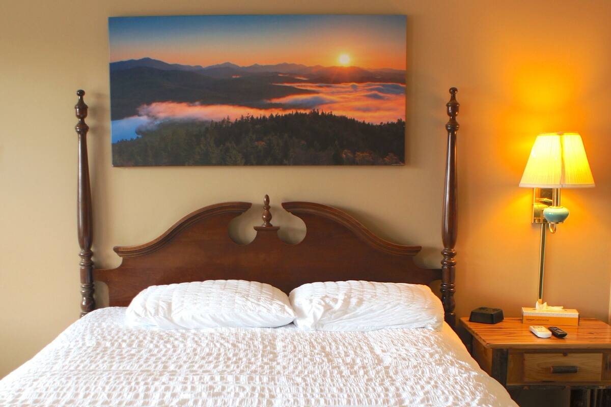 A neatly made bed with a white textured duvet, a wooden headboard, and a painting of a sunset landscape above it.