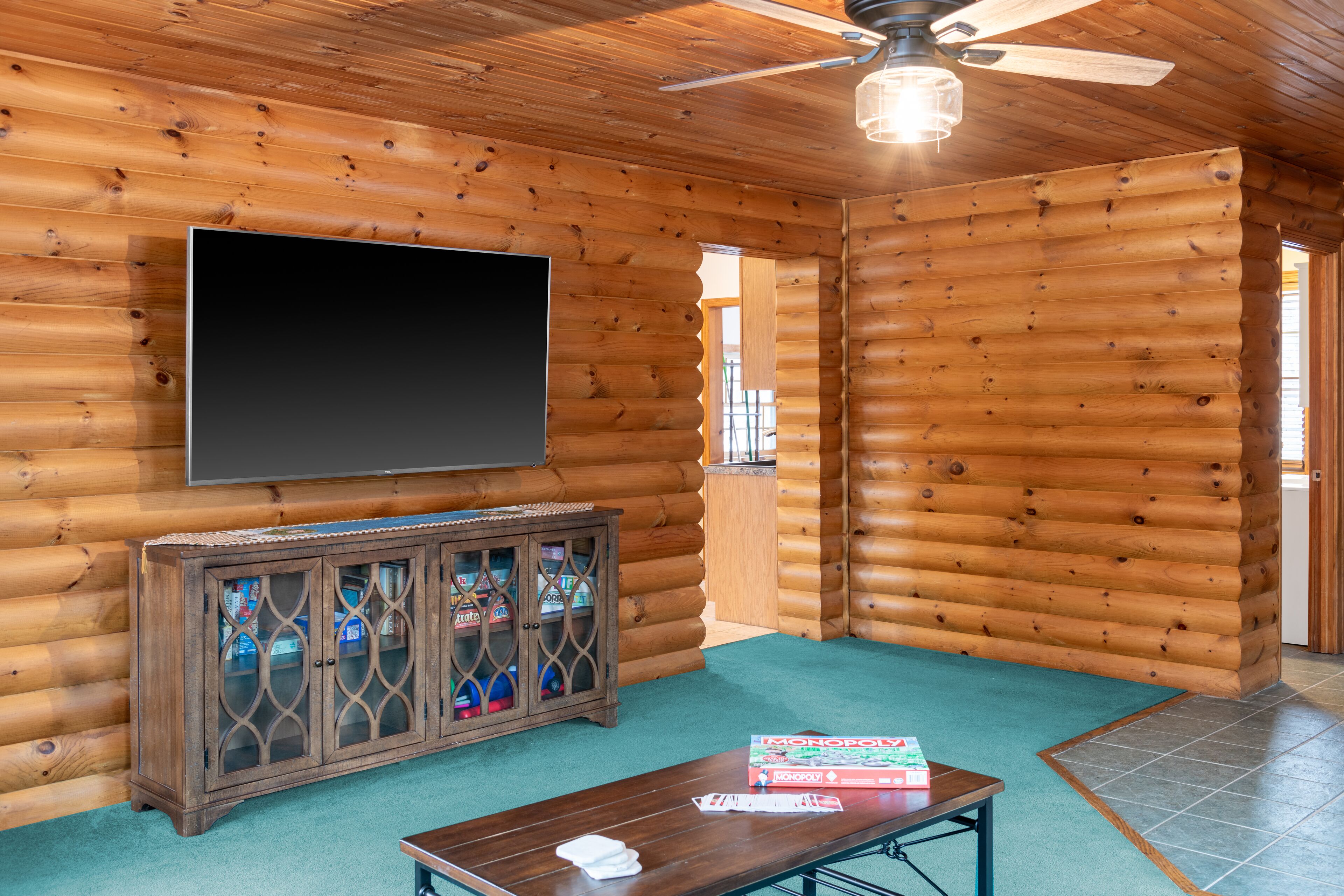 A log cabin interior with a flat-screen TV mounted on the wall, a wooden cabinet with glass doors, a table with a Monopoly box, and a ceiling fan.