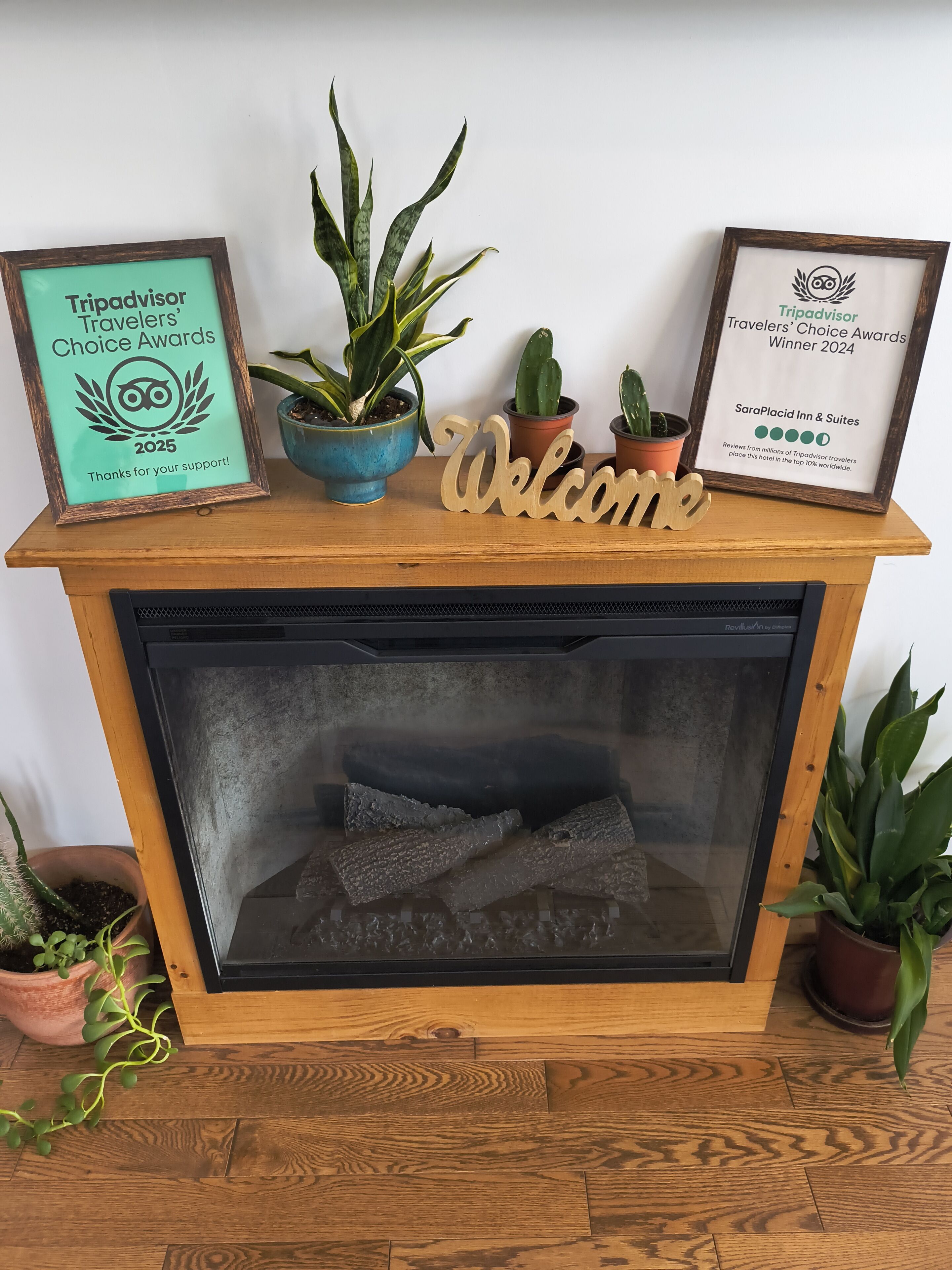 A fireplace with a 'Welcome' sign, framed Tripadvisor awards, and various potted plants.