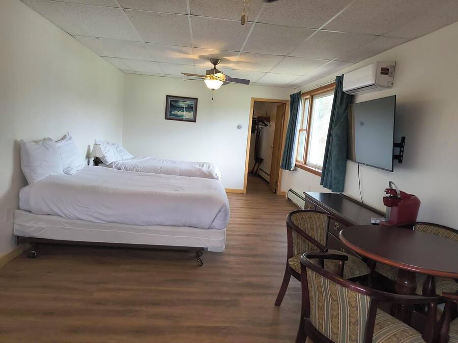 Quarto de hotel com duas camas, um ventilador de teto, uma escrivaninha com cadeira, televisão e uma janela com cortinas.