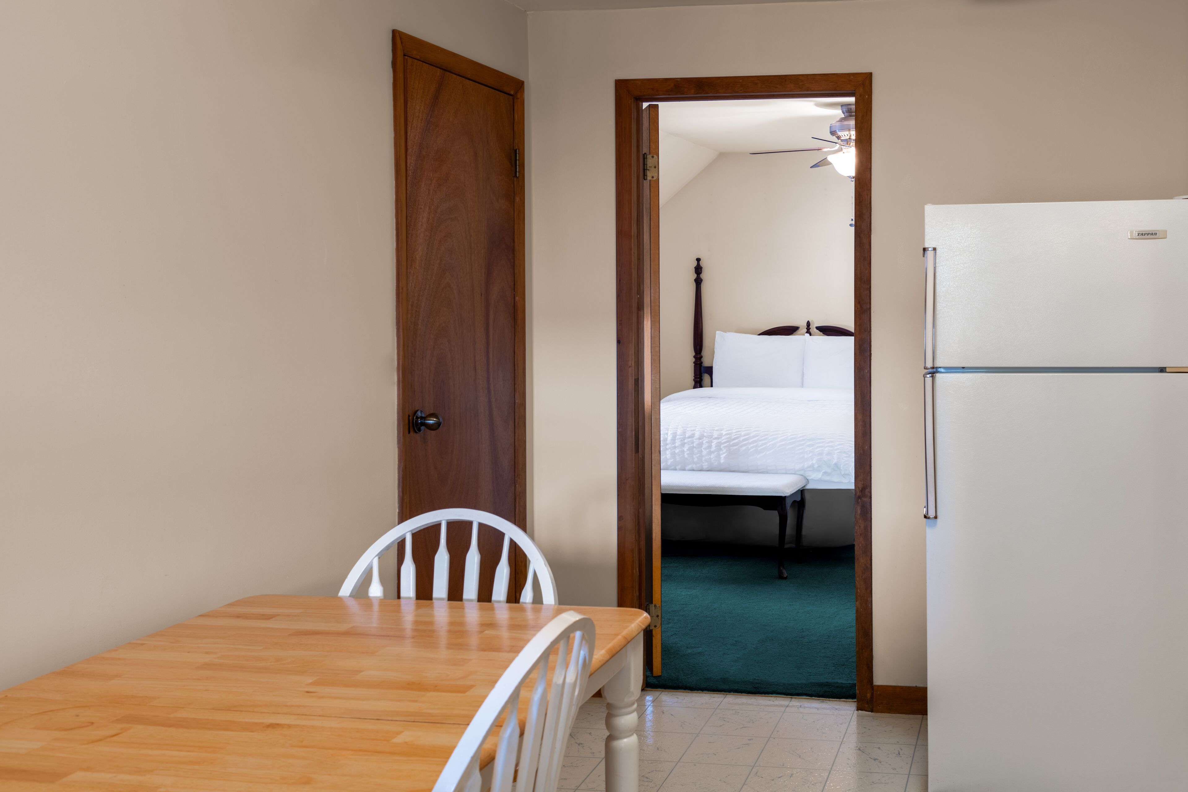 Ein Zimmer mit Holztisch und -stühlen, einem weißen Bett und einem weißen Kühlschrank.