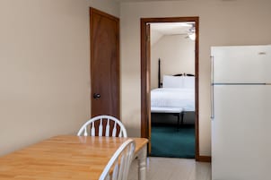 A room with a wooden table and chairs, a white bed, and a white refrigerator.
