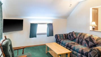 A cozy living room with a sofa, a wooden table, a chair, a TV mounted on the wall, and a window with curtains.