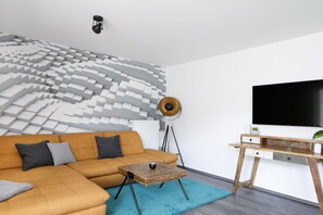 Un salon moderne avec un canapé d’angle, une table basse, un lampadaire et un téléviseur fixé au mur.
