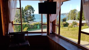 Quarto com uma janela grande que oferece vista para um corpo d'água, vegetação e edifícios ao longe.