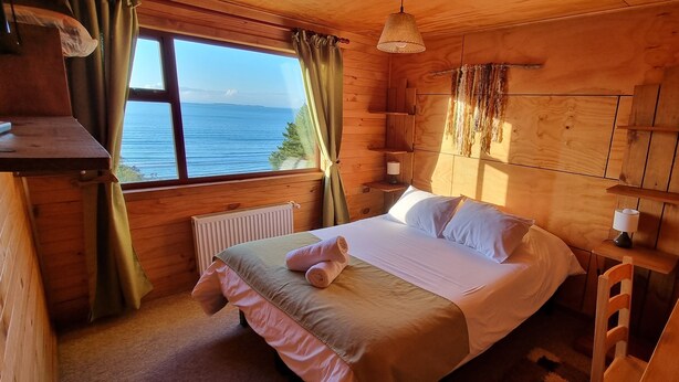 Quarto de cabana de madeira com cama, mesa de cabeceira, janela com cortinas e vista para o mar.