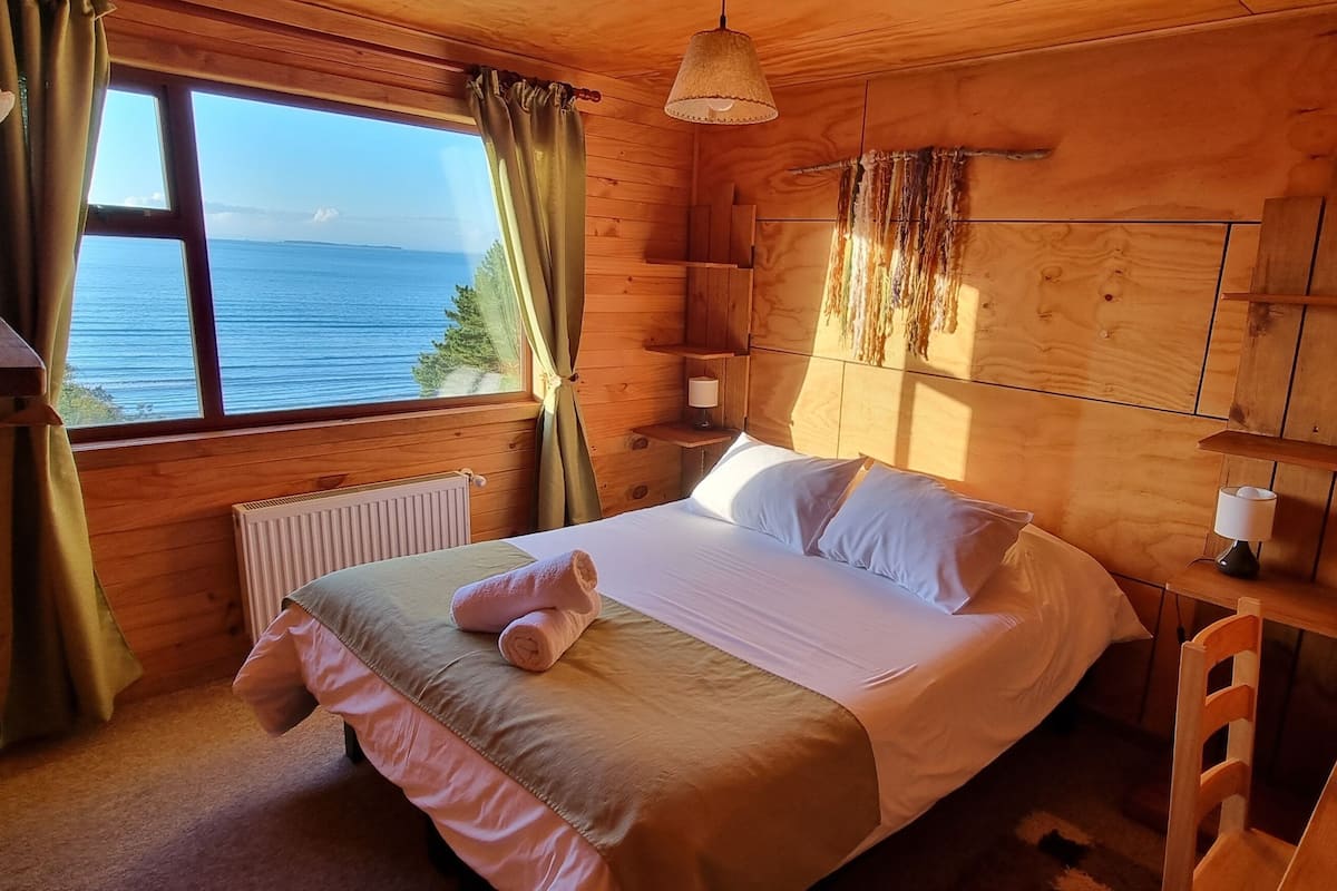 Un dormitorio de cabaña de madera con cama, mesita de noche, ventana con cortinas y vista al mar.