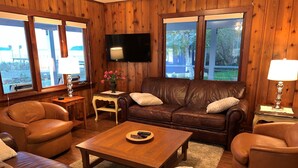 A cozy living room with a brown leather sofa, two leather armchairs, a wooden coffee table, and a TV mounted on the wall. - "Driftwood Island Retreat" Spectacular View, Private Waterfront on James Point. (Vashon)