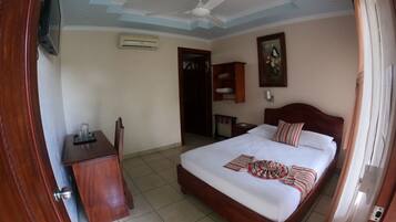 A hotel room with a bed, a desk, a chair, a ceiling fan, an air conditioning unit, and a painting on the wall.