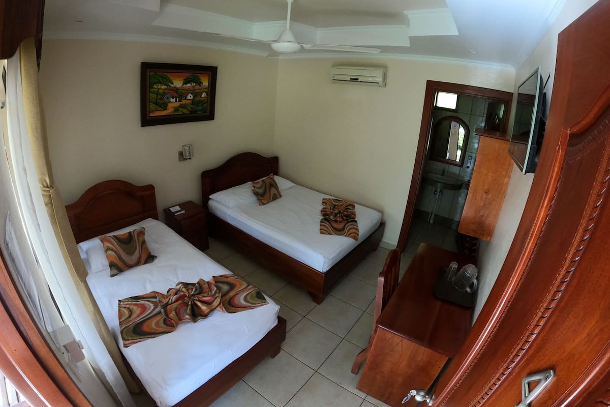 A hotel room with two beds, a ceiling fan, an air conditioning unit, a painting on the wall, and a bathroom visible through an open door.