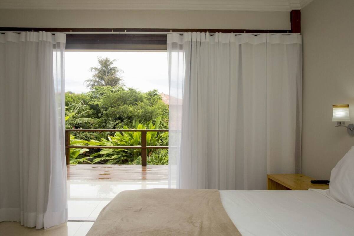 A hotel room with a bed, a nightstand, and a view of lush greenery through a large window with white curtains.