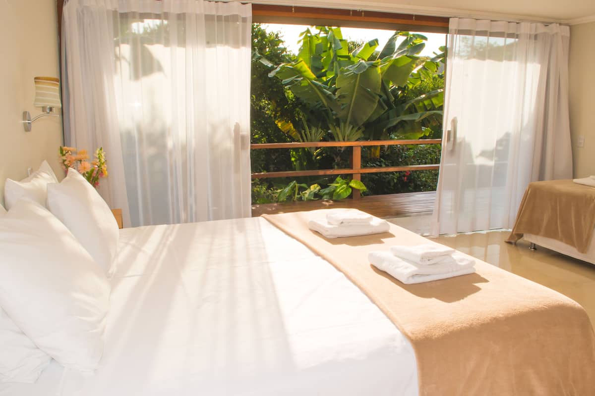 A hotel room with a large bed, a balcony with a view of greenery, and a window with sheer curtains.