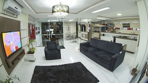 A modern living room with a large flat-screen TV, a black sofa, a chandelier, and a kitchen area in the background. - Hotel Sambakia (Realeza)