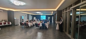 Une salle de conférence avec des tables disposées pour une réunion, un écran de présentation et une personne debout près de la fenêtre.