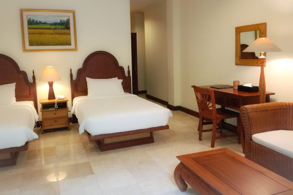 A hotel room with two single beds, a wooden desk with a chair, a small table, and a lamp.