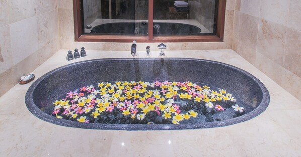 Een groot vrijstaand bad, gevuld met water en kleurrijke bloemen, met een glazen douchecabine erboven.