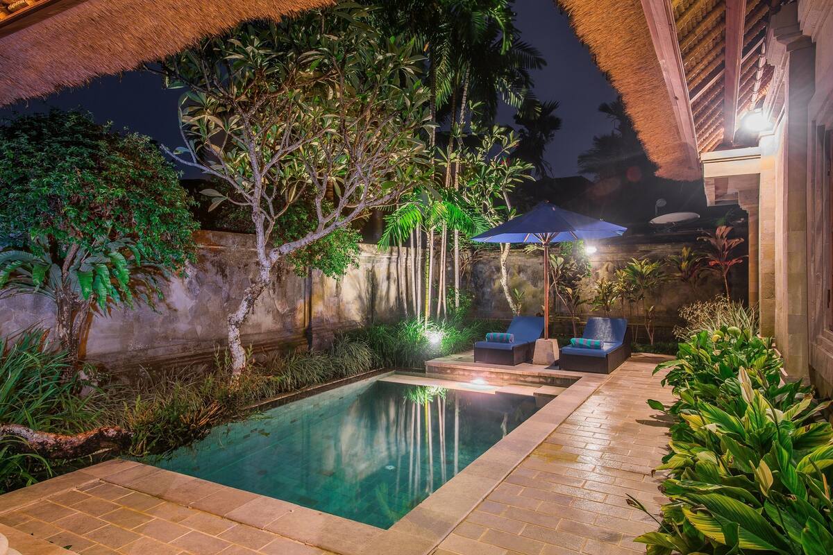 A swimming pool with lounge chairs and umbrellas, surrounded by lush greenery and stone walls.