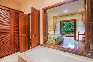 Un baño con bañera, puertas de madera y un espejo grande que refleja un dormitorio con cama y cortinas.