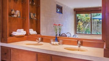 Un baño con un amplio tocador de madera, dos lavamanos y un espejo grande. Hay estantes con toallas y artículos de aseo, y una ventana con vistas a la vegetación.