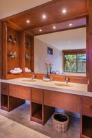 A bathroom with a large wooden vanity, two sinks, and a large mirror. There are shelves with towels and toiletries, and a window with a view of greenery.