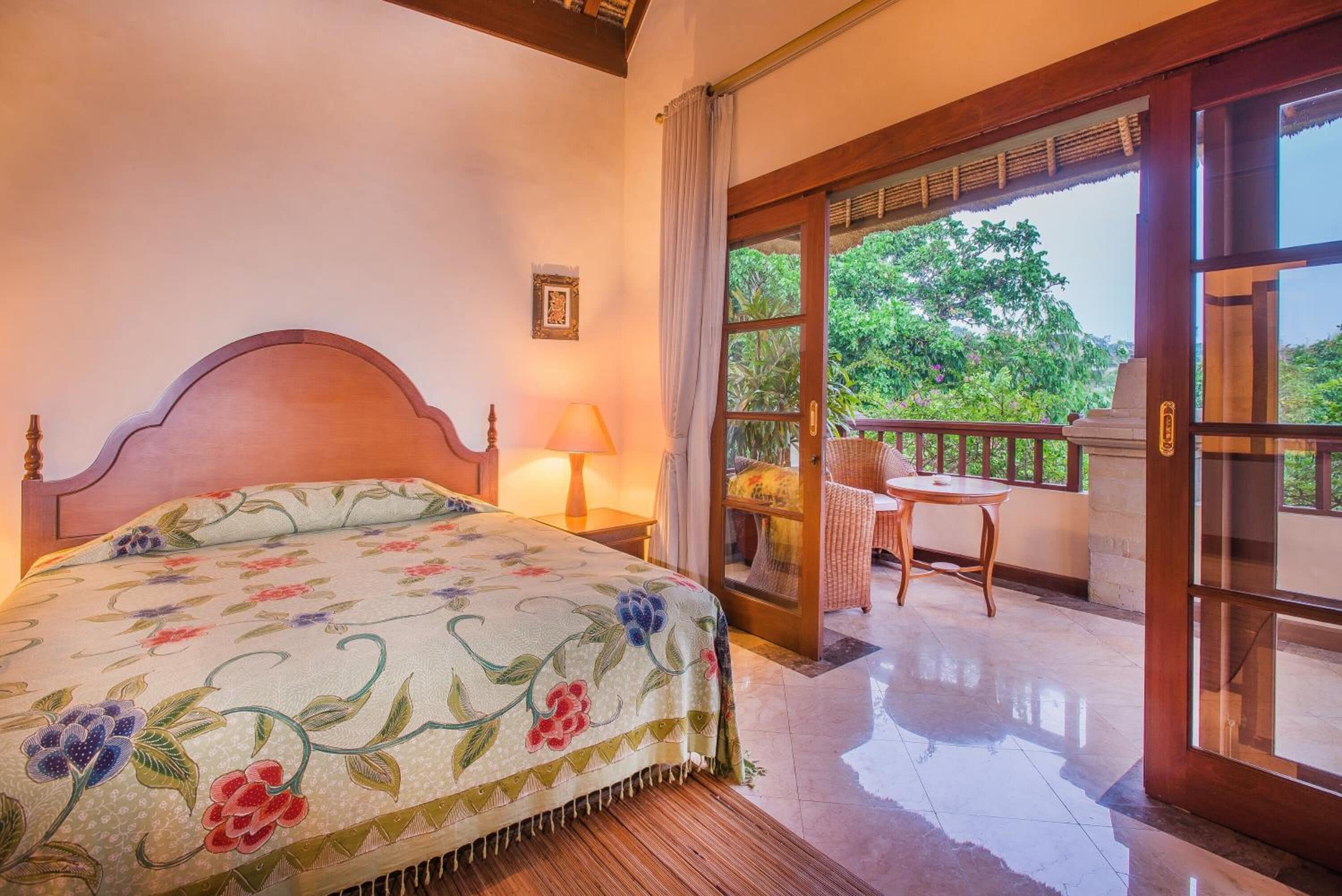 A bedroom with a large bed, a nightstand, a lamp, and a balcony with a chair and table.