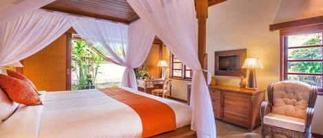 A four-poster bed with white and orange bedding, a wooden desk, a wicker armchair, and a TV on the wall.